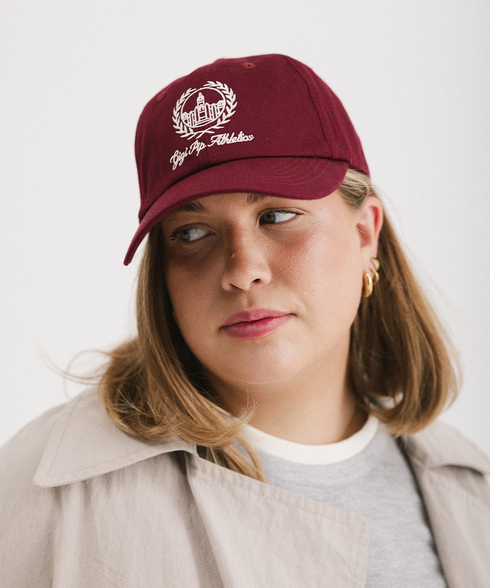 Woman wearing a burgundy ball cap with a white logo and text and a grey t-shirt with a beige shirt, looking sideways against a plain background  #color_burgundy