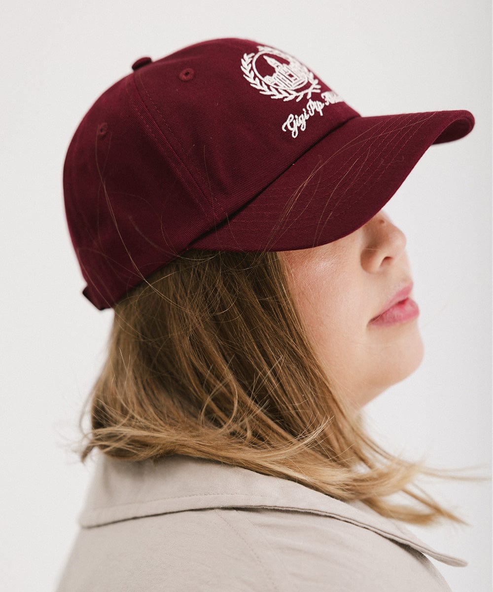 Woman wearing a burgundy ball cap with a white logo and text facing sideways against a plain background  #color_burgundy