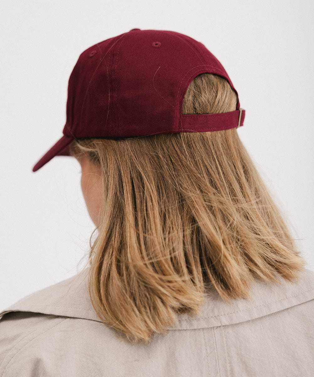 Woman wearing a burgundy ball cap and a beige shirt, facing behind against a plain background  #color_burgundy