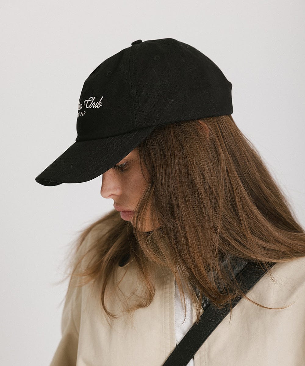 Woman wearing a black cap with white 'Athletics Club' text and a beige jacket, facing sideways against a plain background  #color_black