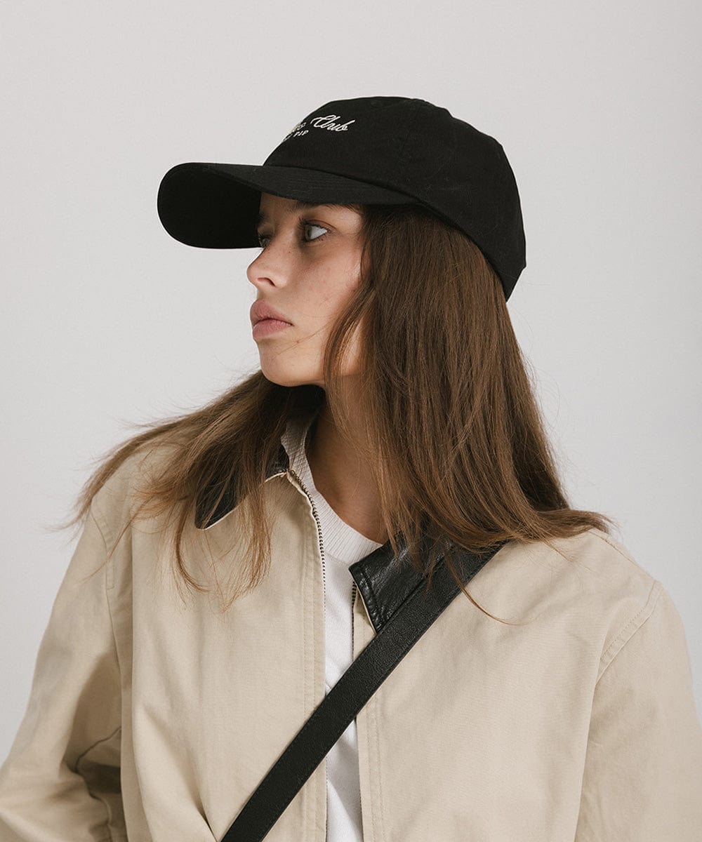 Woman wearing a black cap with white 'Athletics Club' text and a beige jacket, posing sideways against a plain background  #color_black