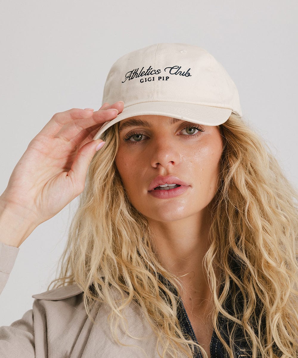 Woman wearing a cream cap with black 'Athletics Club' text and a beige shirt against a plain background #color_cream