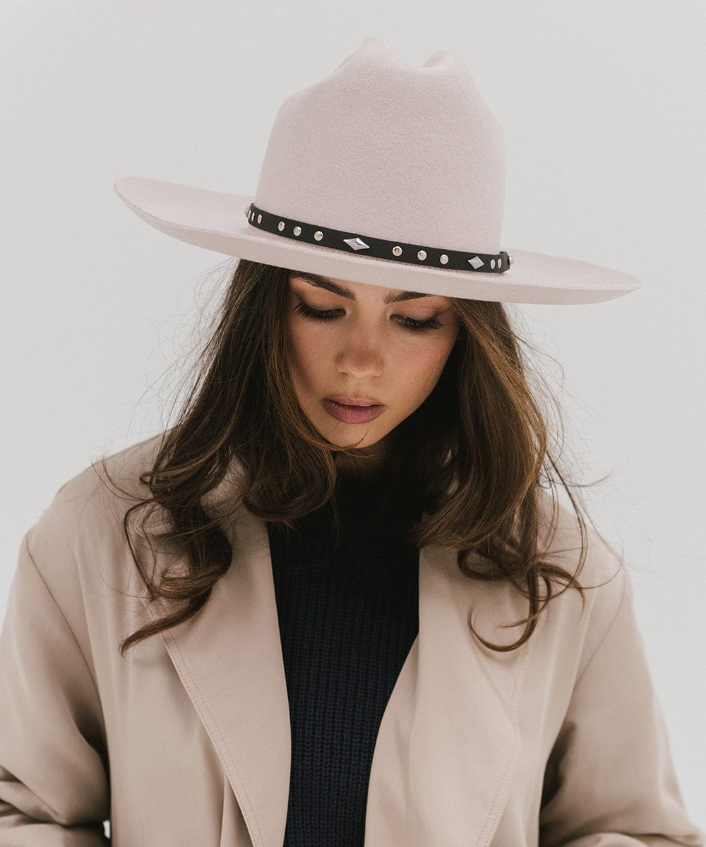 Woman wearing a white hat with a black studded leather hat band and a beige coat against a plain background #color_black