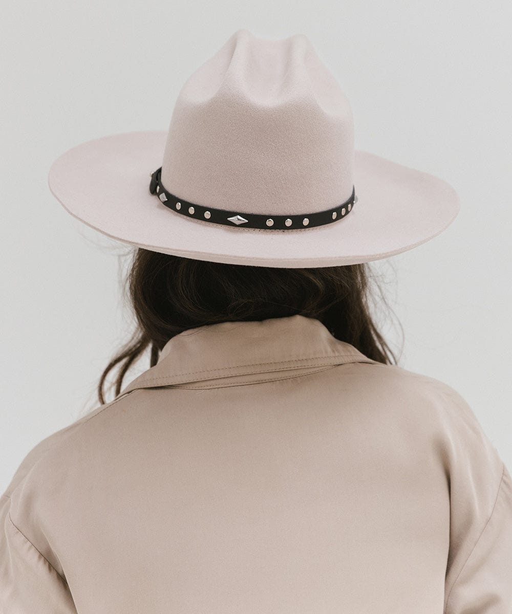 Person wearing a white hat with a black studded leather hat band and a beige coat against a plain background #color_black