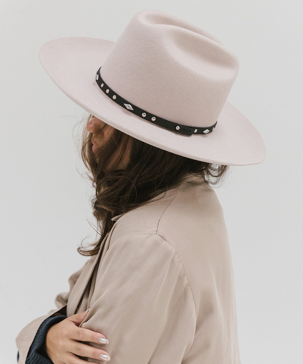 Woman wearing a white hat with a black studded leather hat band and a beige coat, facing sideways against a plain background #color_black