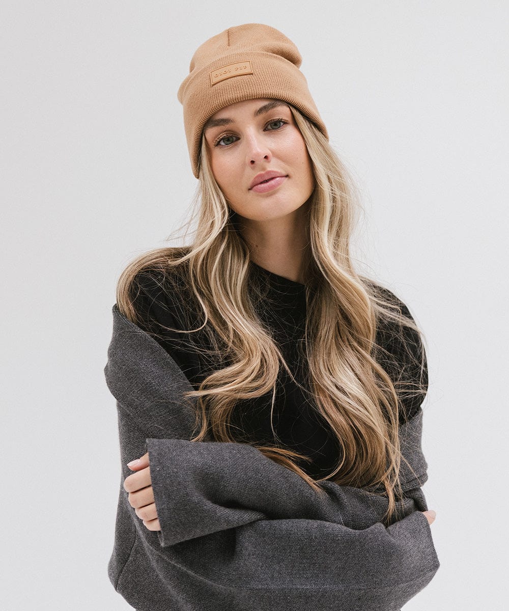 Woman wearing a latte colored knit beanie and a black t-shirt with a grey sweater against a plain background #color_latte