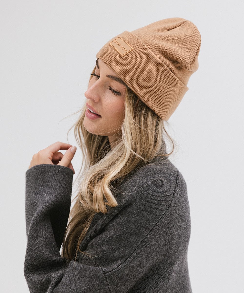 Woman wearing a latte colored knit beanie and a grey sweater, facing sideways against a plain background #color_latte