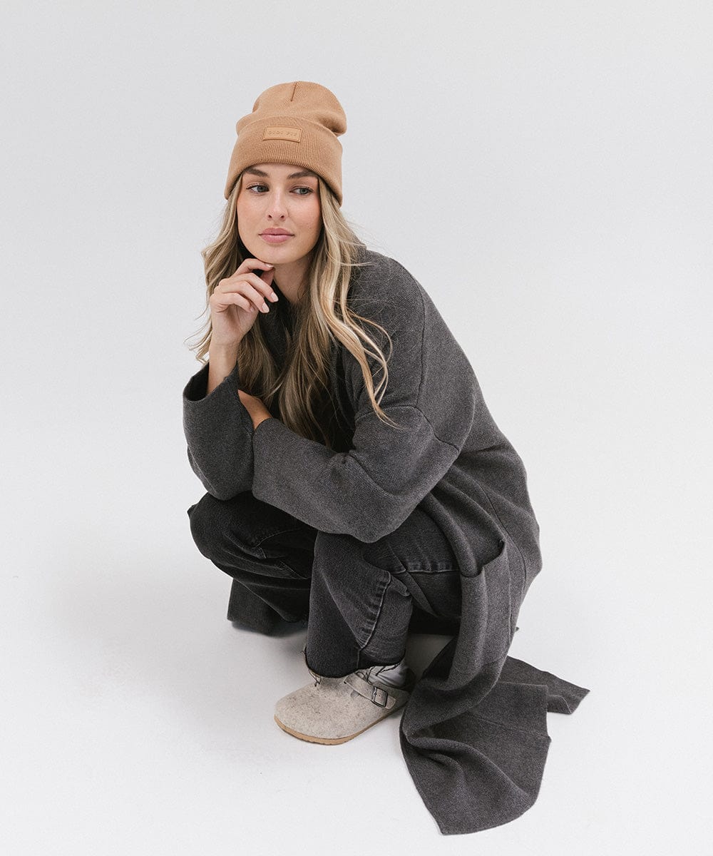 Woman wearing a latte colored knit beanie, a grey coat and black jeans, sitting against a plain background #color_latte