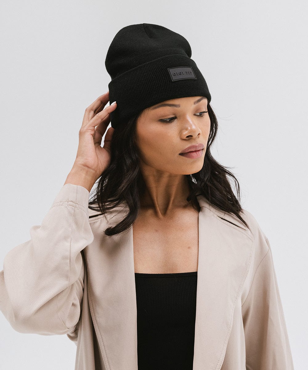 Woman wearing a black knit beanie and a black top with a beige coat against a plain background #color_black