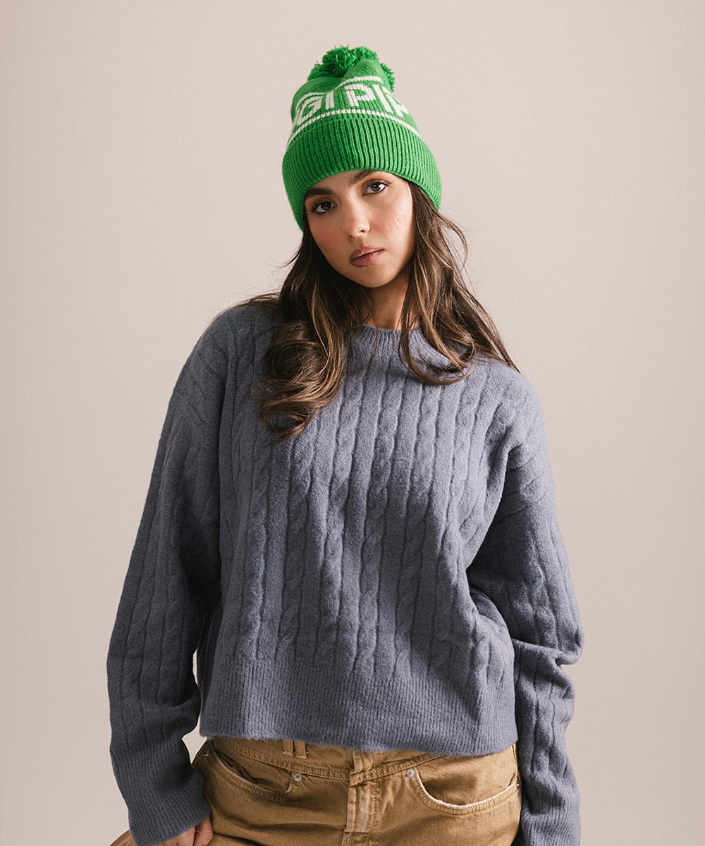 Woman wearing an evergreen colored knit beanie with pom-pom and 'GIGI PIP' branding and a blue sweater against a plain background 