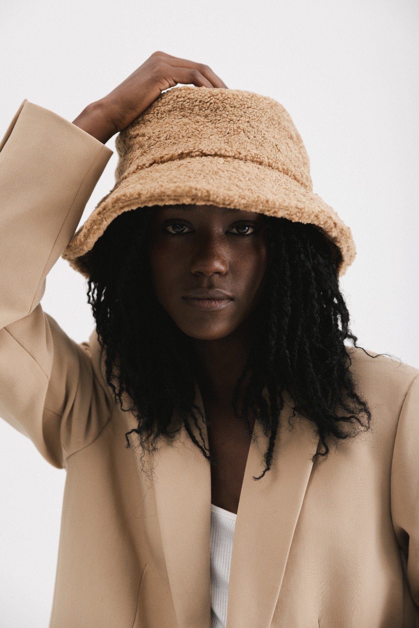 Woman wearing a camel colored sherpa bucket hat and a beige coat  against a plain background #color_camel