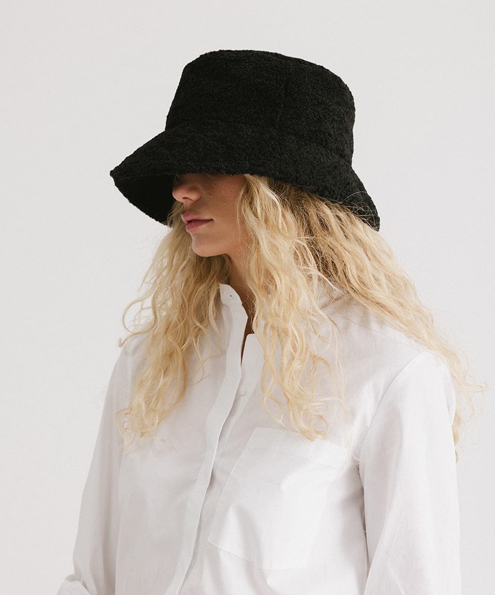 Woman wearing a black sherpa bucket hat and a white shirt, facing sideways against a plain background #color_black