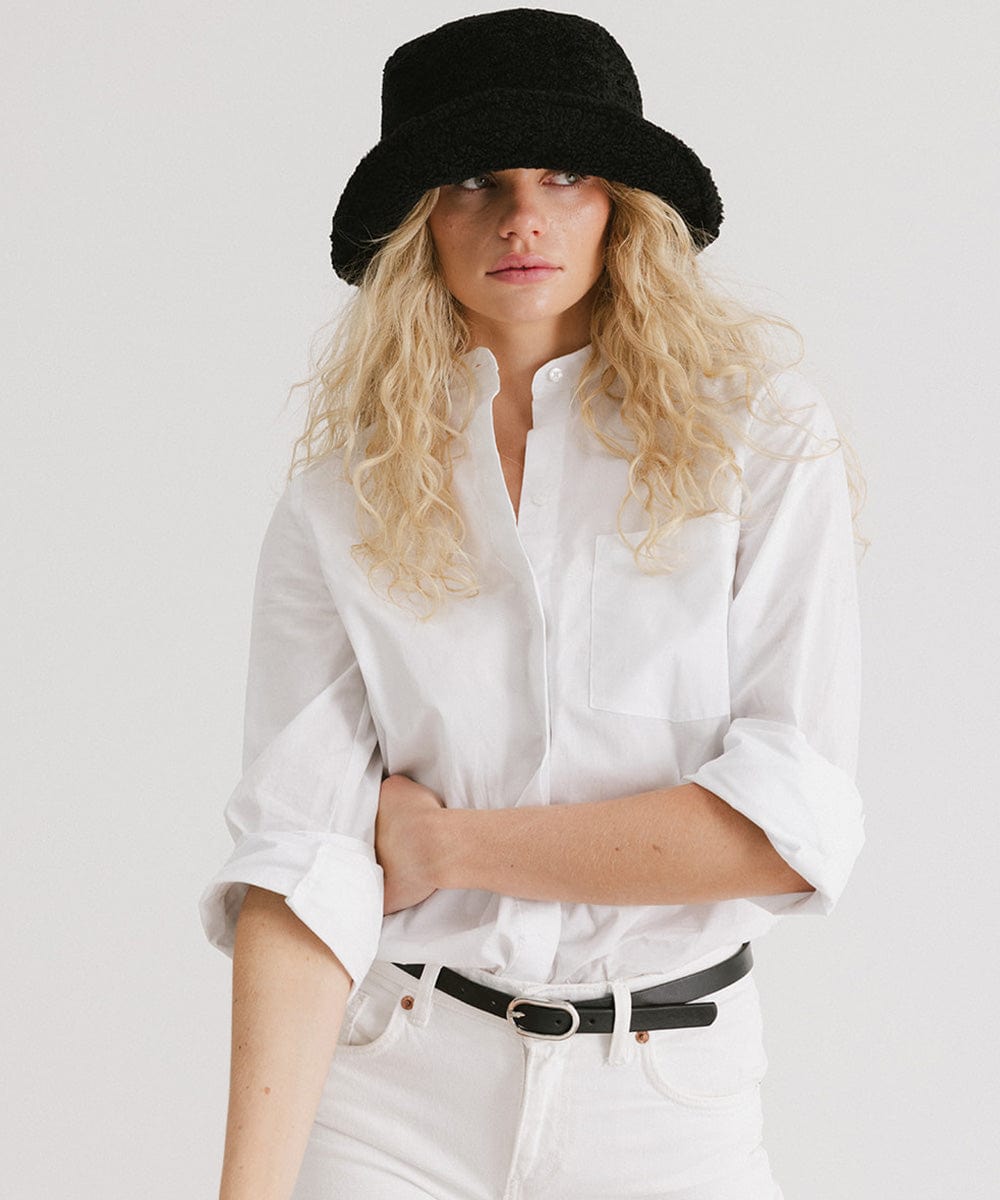 Woman wearing a black sherpa bucket hat, a white shirt, black belt and white pants against a plain background #color_black