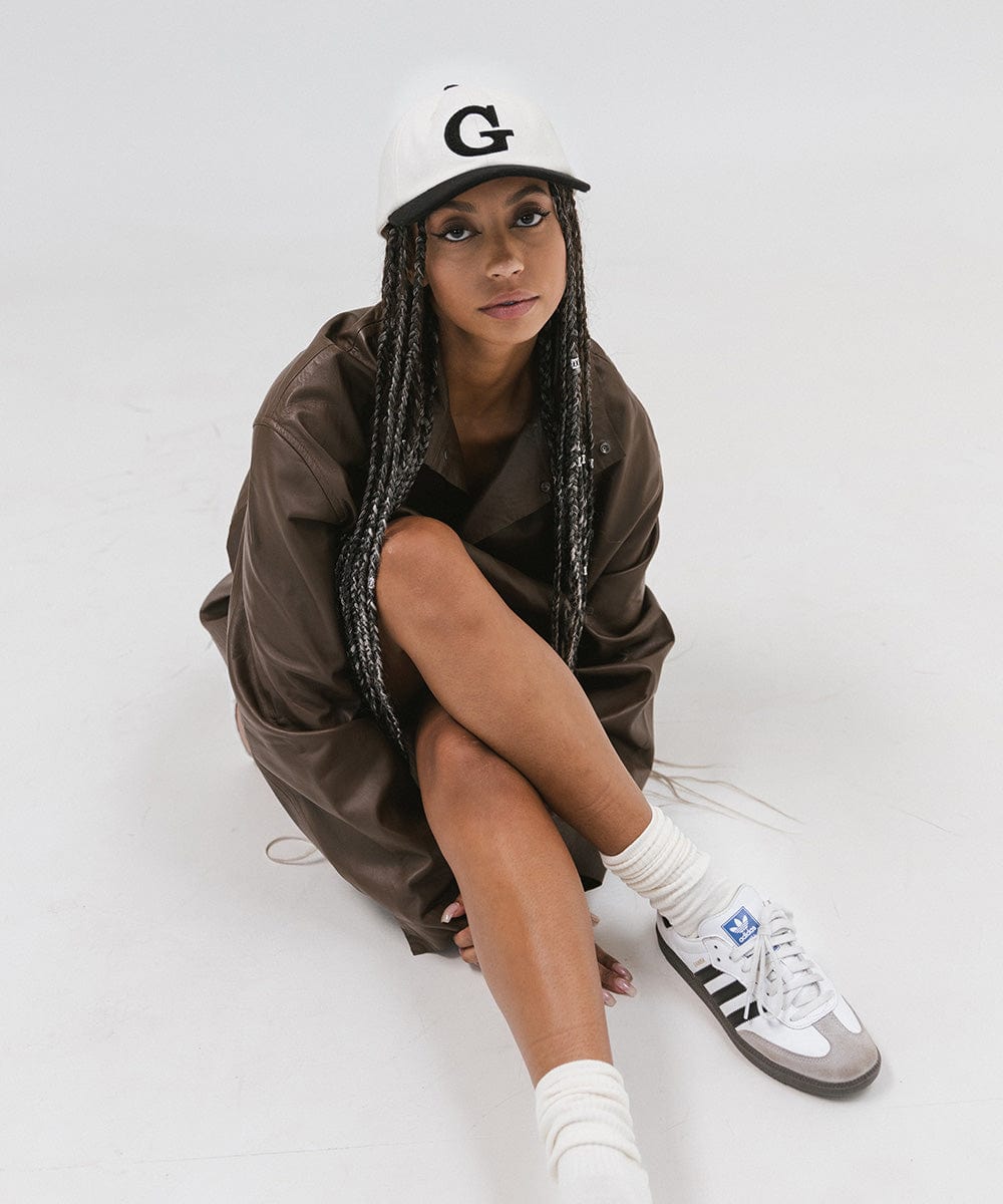 Woman wearing a cream and black baseball cap with the letter 'G', a brown jacket and white shoes, sitting against a plain background  #color_cream-black