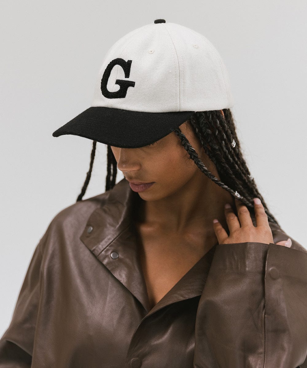 Woman wearing a cream and black baseball cap with the letter 'G' and a brown jacket, posing against a plain background  #color_cream-black