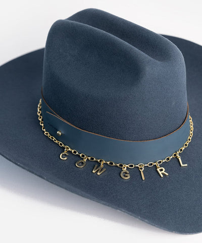 A blue cowboy hat with a hat band and a gold chain with letter hat charms making up the word 'COWGIRL' on a plain background #color_gold
