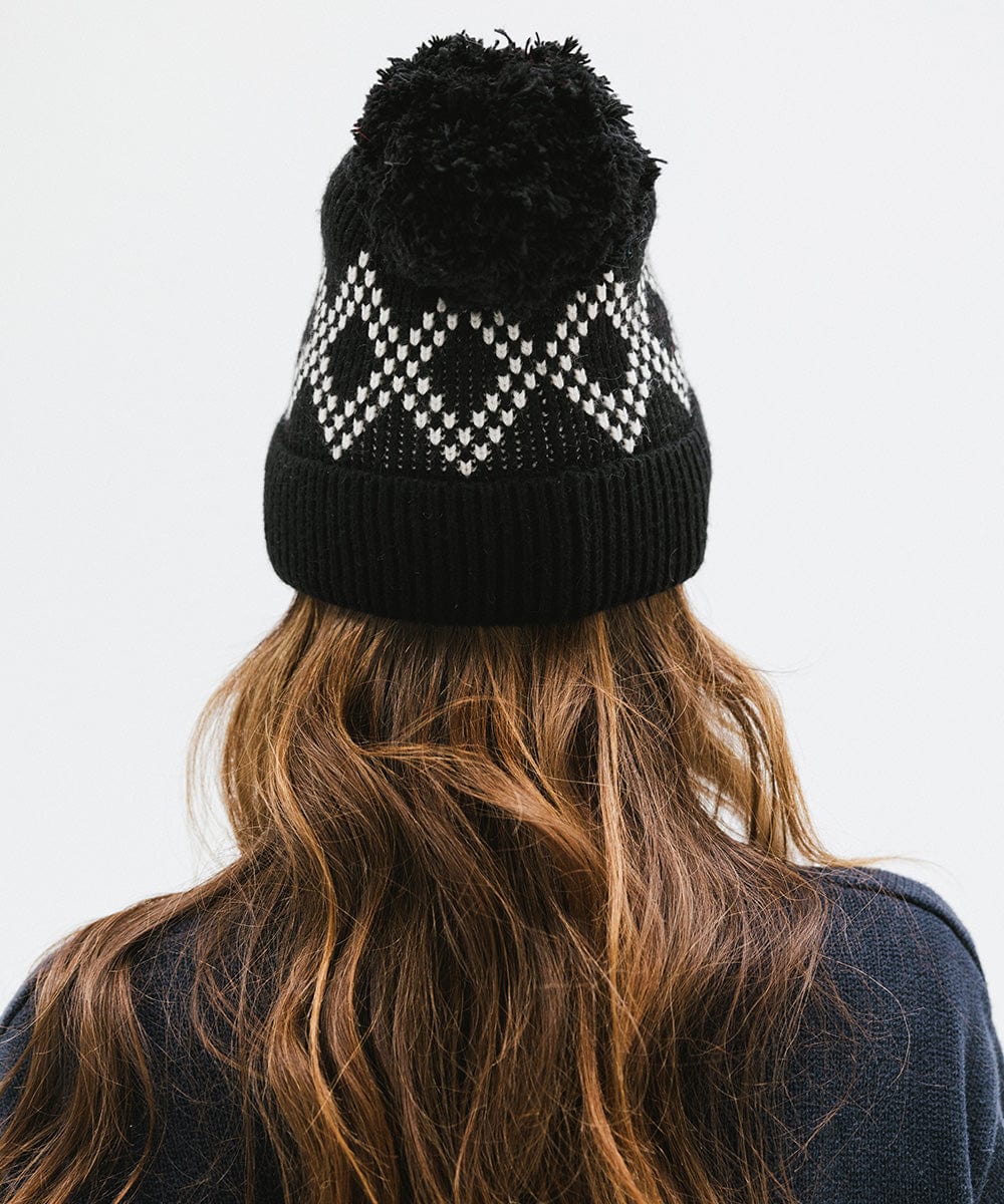 Woman wearing a black knit beanie with a pom-pom and a dark blue sweater, facing behind against a plain background #color_black