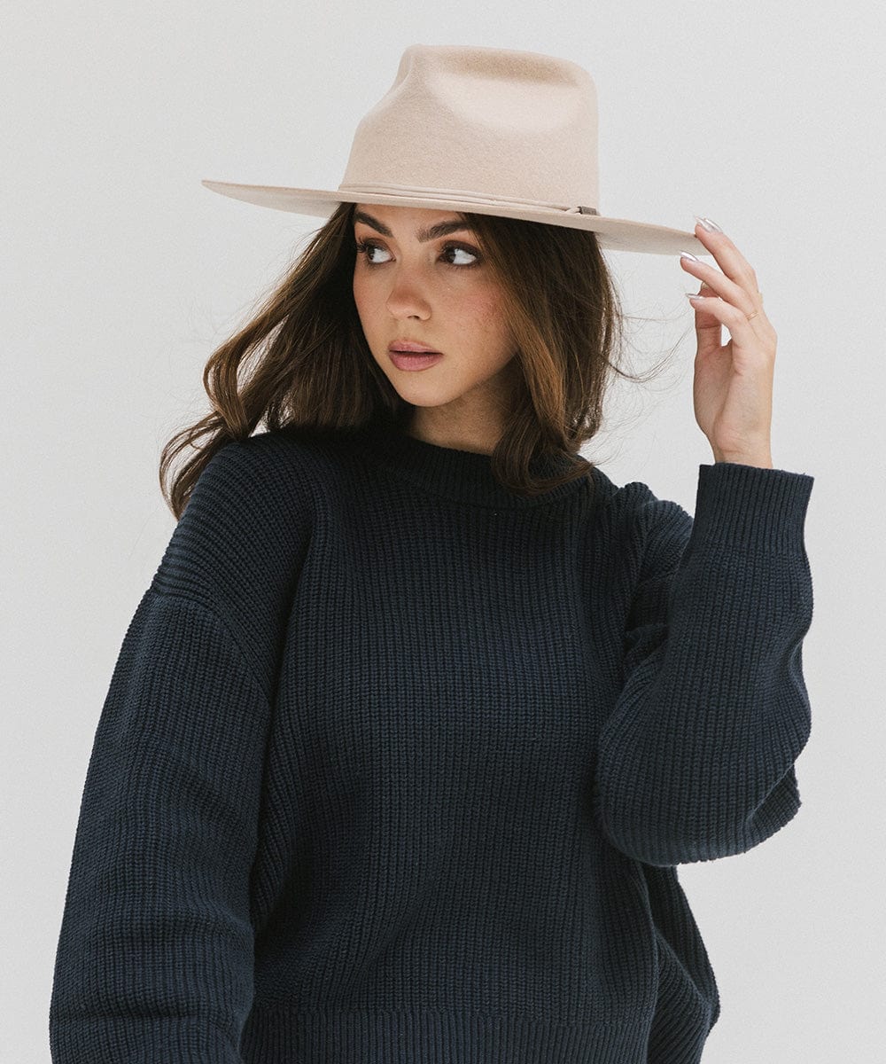 Woman wearing a cream fedora hat with a hat band and a dark blue sweatshirt, looking sideways against a plain background #color_cream