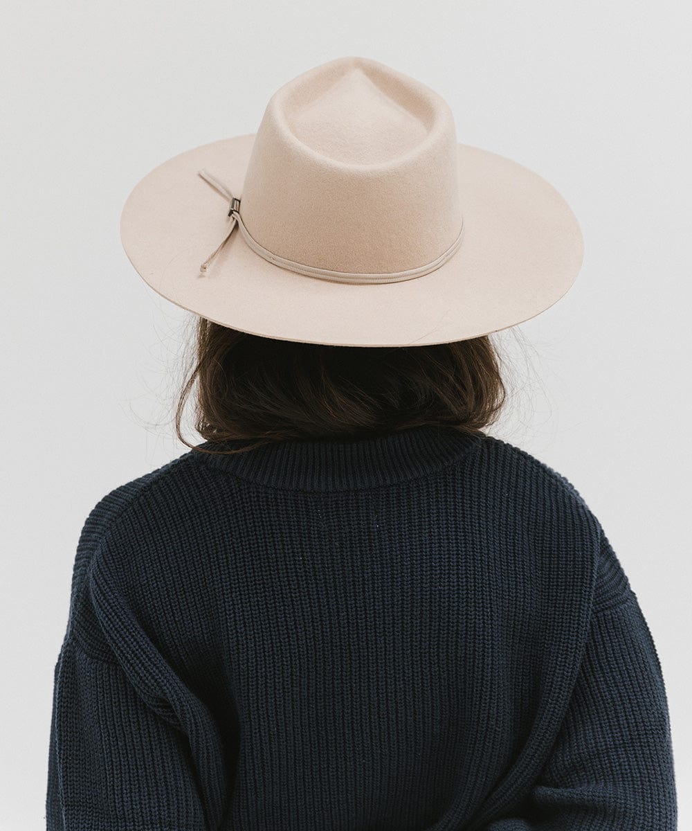 Woman wearing a cream fedora hat with a hat band and a dark blue sweatshirt, facing behind against a plain background #color_cream