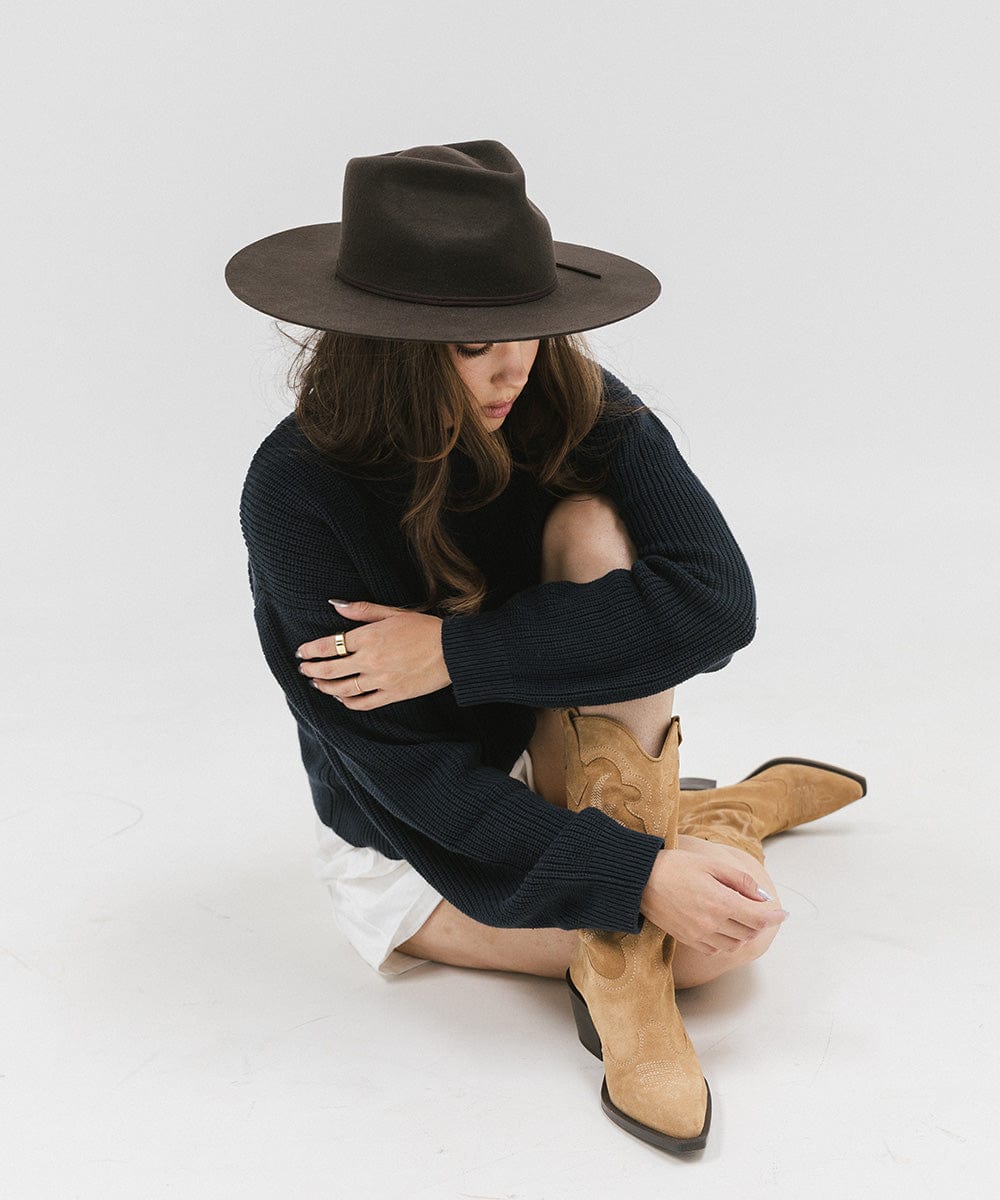 Woman wearing a dark brown fedora hat, a dark blue sweatshirt, white shorts and brown boots, sitting against a plain background #color_dark brown