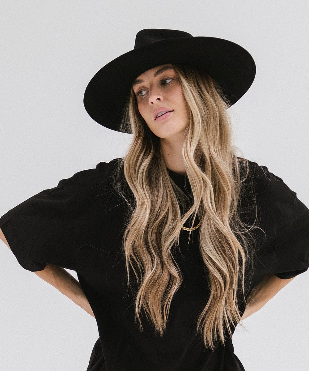 Woman with long hair wearing a black fedora hat with a hat band and a black t-shirt, posing against a plain background #color_black