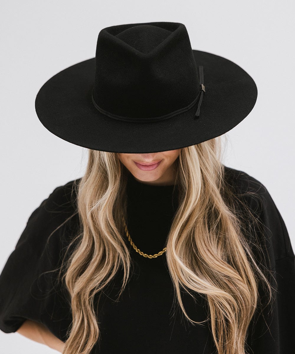 Woman with long hair wearing a black fedora hat with a hat band, a gold chain and a black t-shirt, looking down against a plain background #color_black