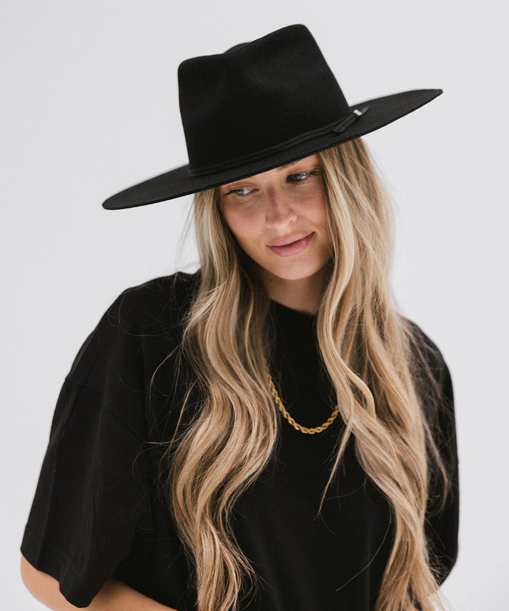 Woman with long hair wearing a black fedora hat with a hat band and a black t-shirt against a plain background #color_black