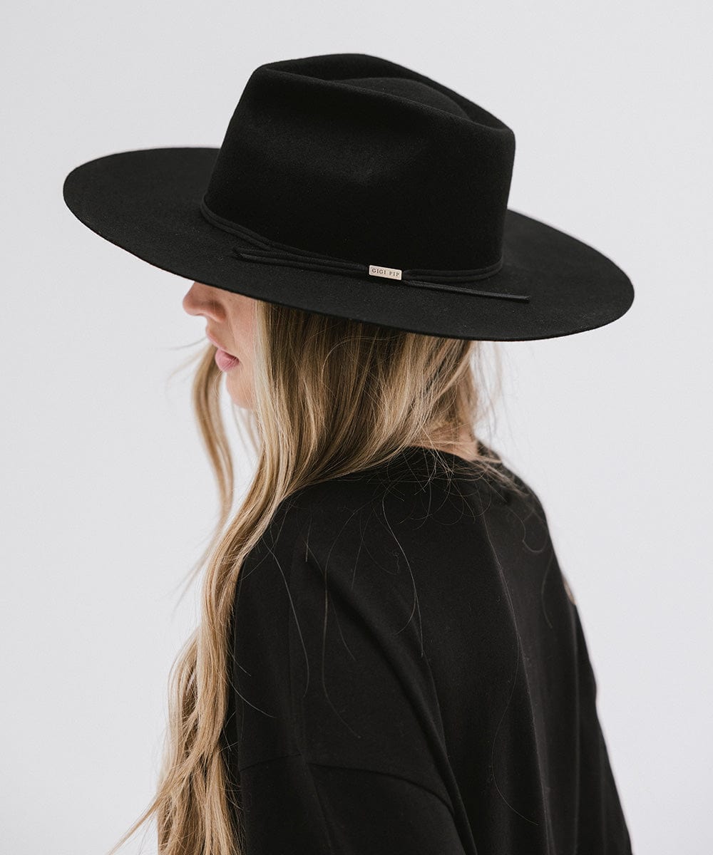 Woman with long hair wearing a black fedora hat with a hat band and a black t-shirt, facing sideways against a plain background #color_black