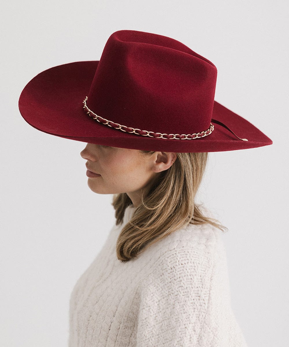 Woman wearing a burgundy colored felt hat with a metallic chain detail on the band and a white sweater, facing sideways against a plain background  #color_burgundy