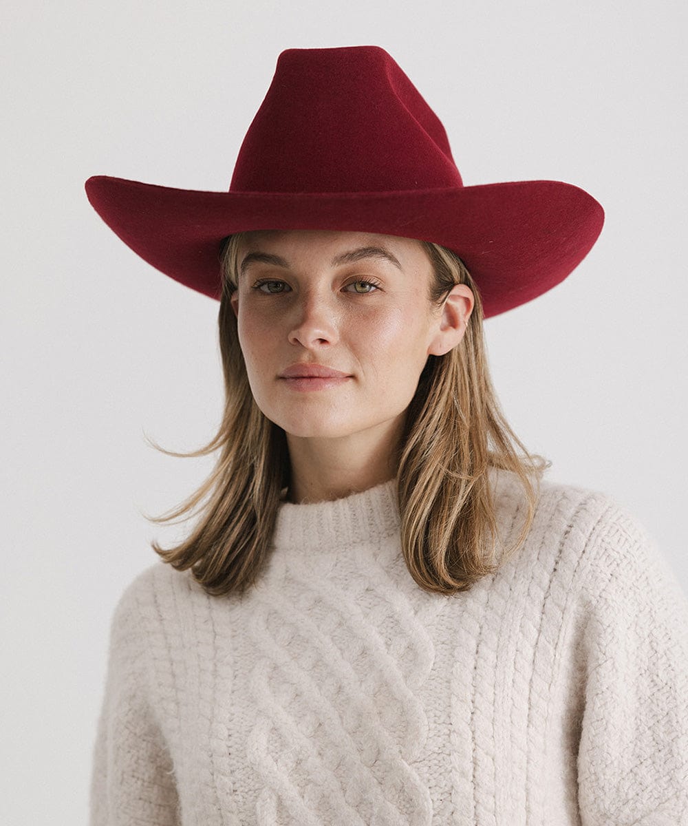 Woman wearing a burgundy colored felt hat with a metallic chain detail on the band and a white sweater against a plain background   #color_burgundy