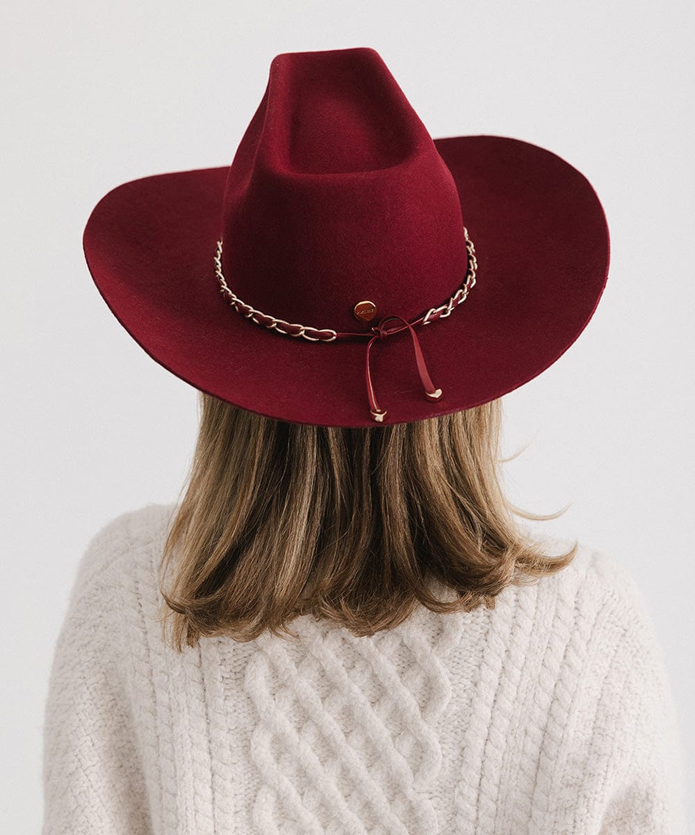 Person wearing a burgundy colored felt hat with a metallic chain detail on the band and a white sweater against a plain background   #color_burgundy
