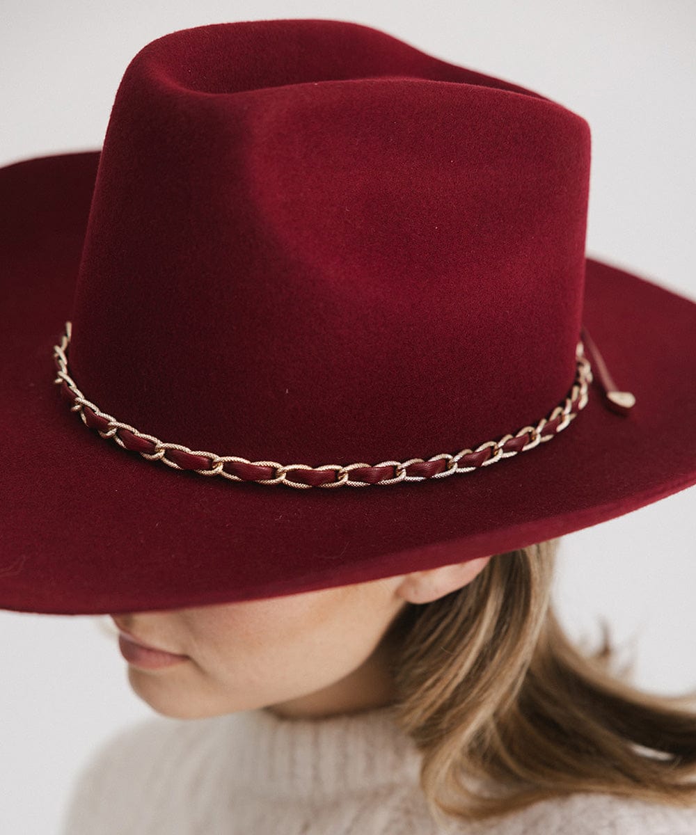 Person wearing a burgundy colored felt hat with a metallic chain detail on the band against a plain background  #color_burgundy