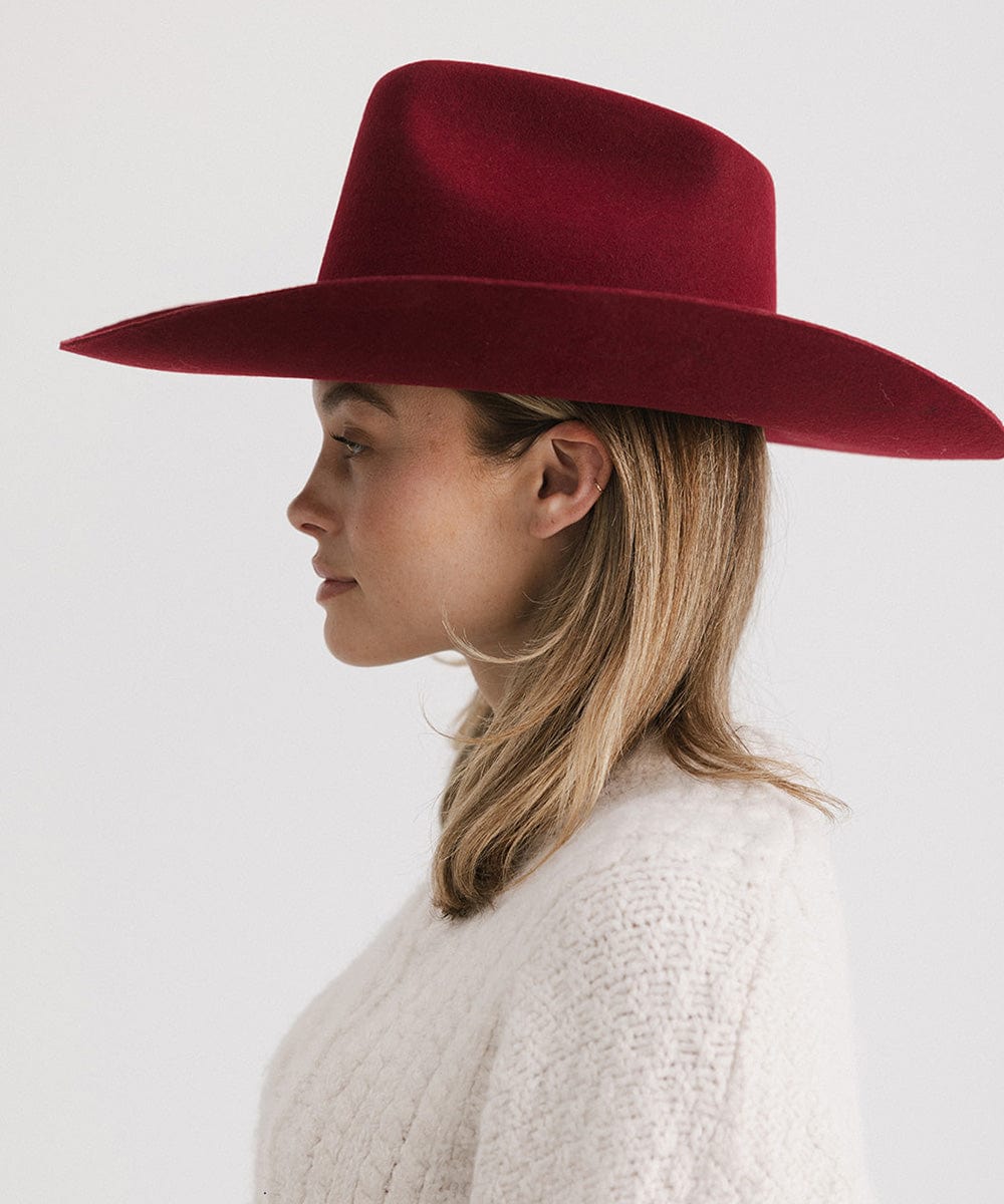 Woman wearing a burgundy colored felt hat and a white sweater, facing sideways against a plain background   #color_burgundy