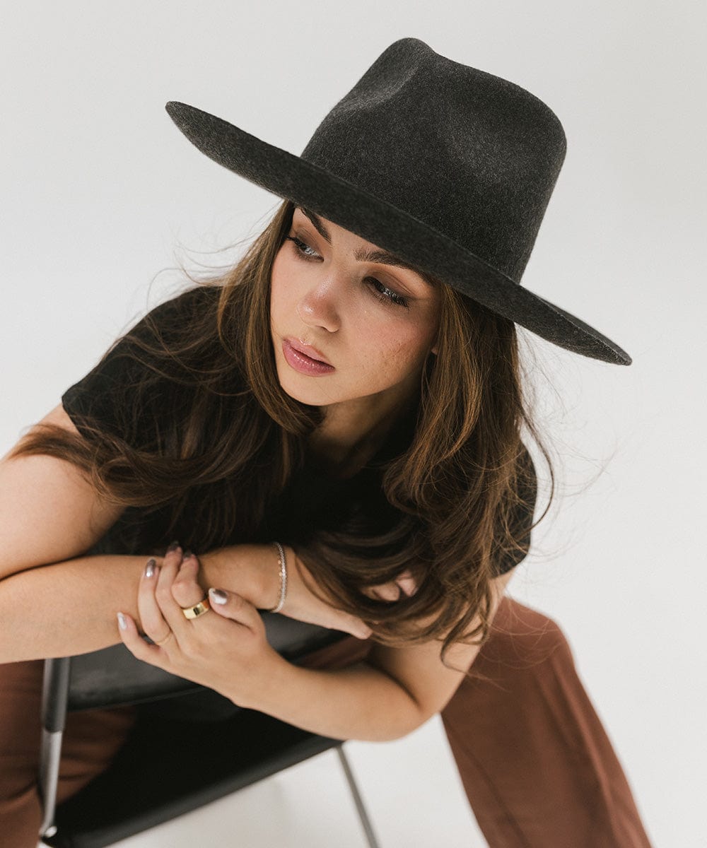 Woman wearing a mix charcoal  colored Rowan Wide Brim Fedora Hat, a black t-shirt and brown pants, sitting on a chair against a plain background  #color_mix charcoal