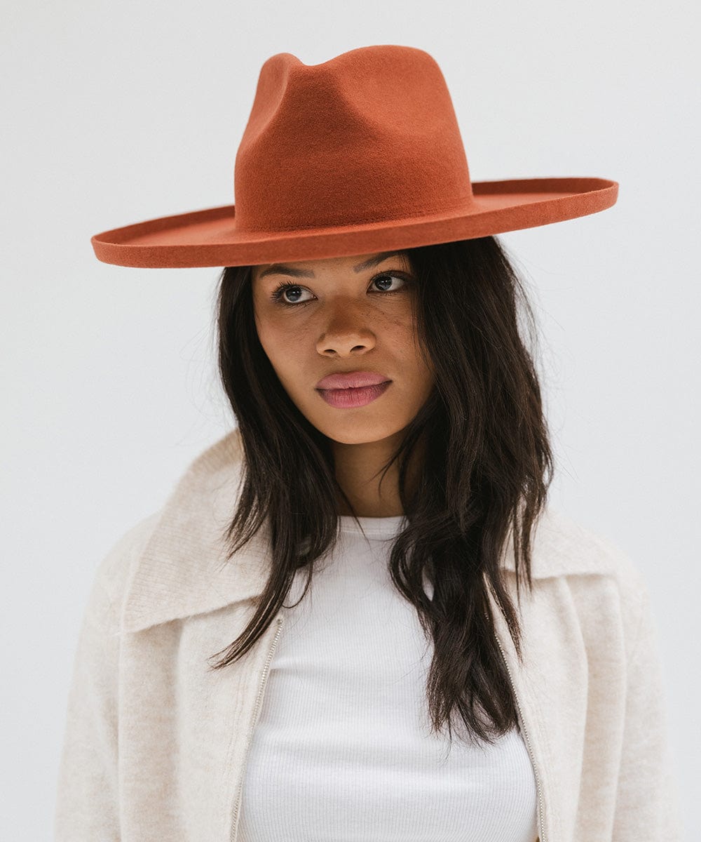 Woman wearing a clay colored pencil brim fedora hat with a wide brim and a white top with a cream jacket against a white background #color_clay