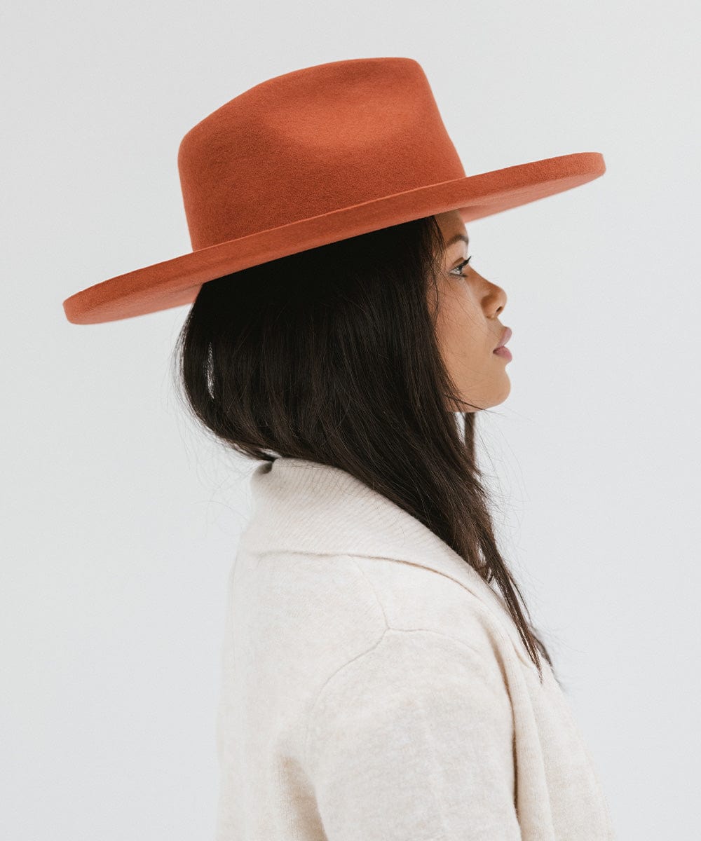 Woman wearing a clay colored pencil brim fedora hat with a wide brim and a cream jacket, facing sideways against a white background #color_clay
