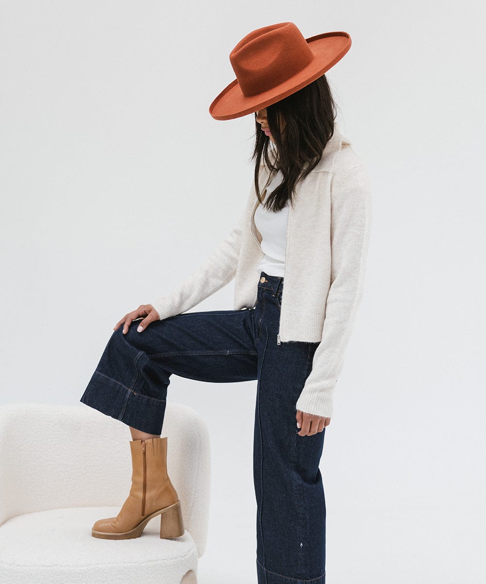 Woman wearing a clay colored pencil brim fedora hat with a wide brim, a cream jacket, dark blue jeans and brown boots, posing against a white background #color_clay