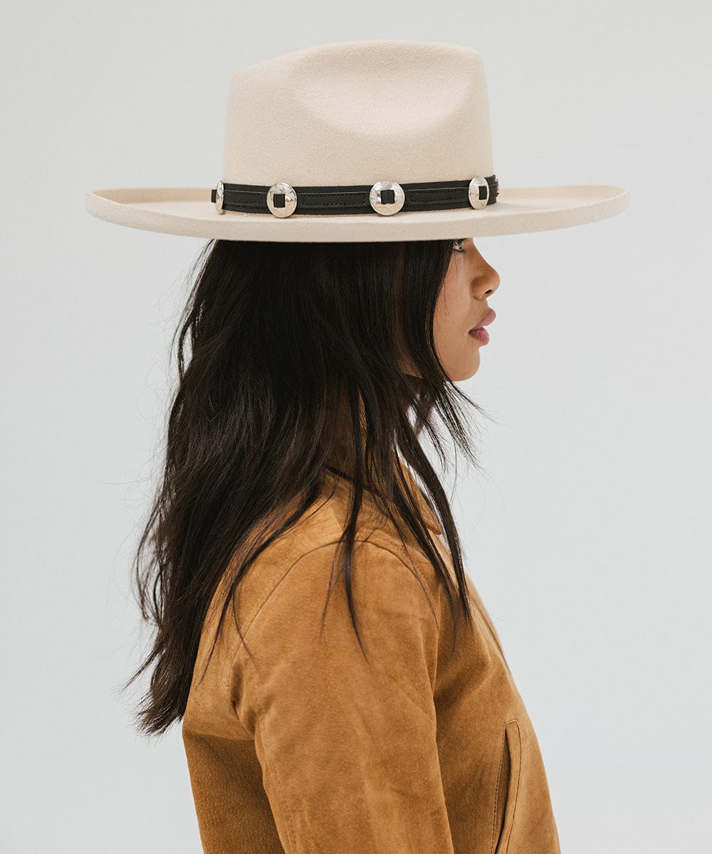 Woman wearing an off white colored pencil brim fedora hat with a wide brim and a hat band, and a brown jacket, facing sideways against a white background  #color_off white