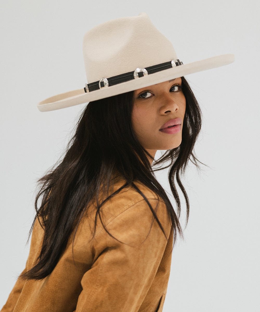 Woman wearing an off white colored pencil brim fedora hat with a wide brim and a hat band, and a brown jacket, posing against a white background  #color_off white
