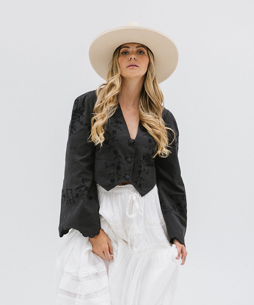 Woman wearing a cream colored pencil brim fedora hat with a wide brim, a black printed shirt, and white pants against a white background #color_cream