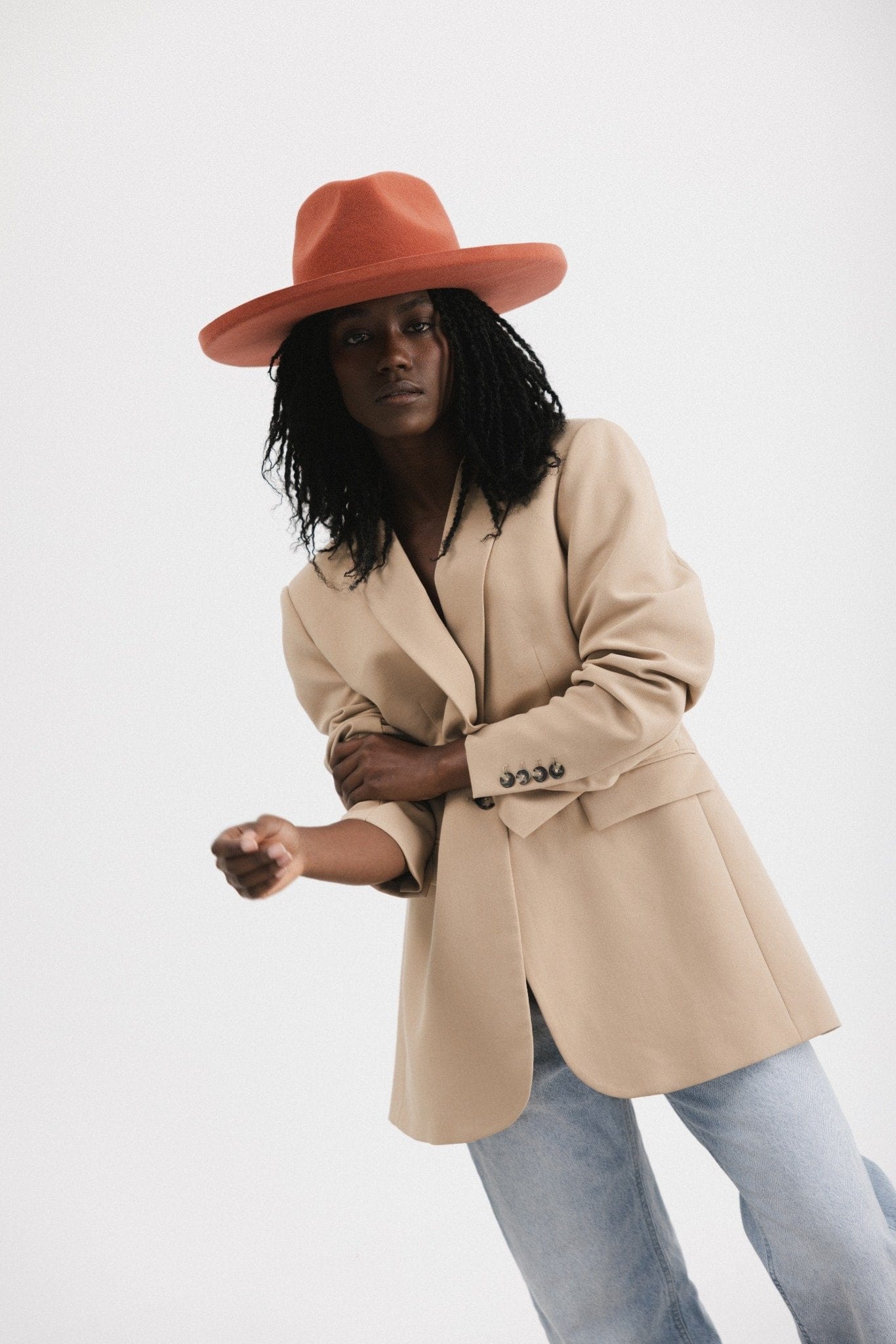Woman wearing a clay colored pencil brim fedora hat with a wide brim, a light brown blazer and light blue jeans against a white background #color_clay