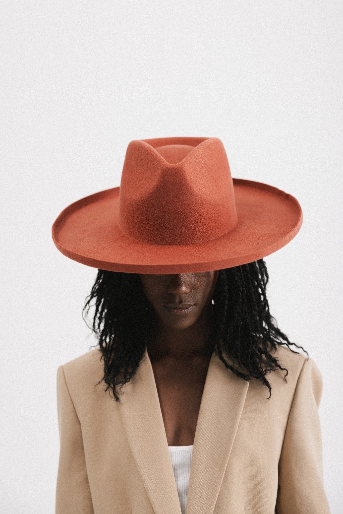 Woman wearing a clay colored pencil brim fedora hat with a wide brim and a light brown blazer, looking down against a white background #color_clay