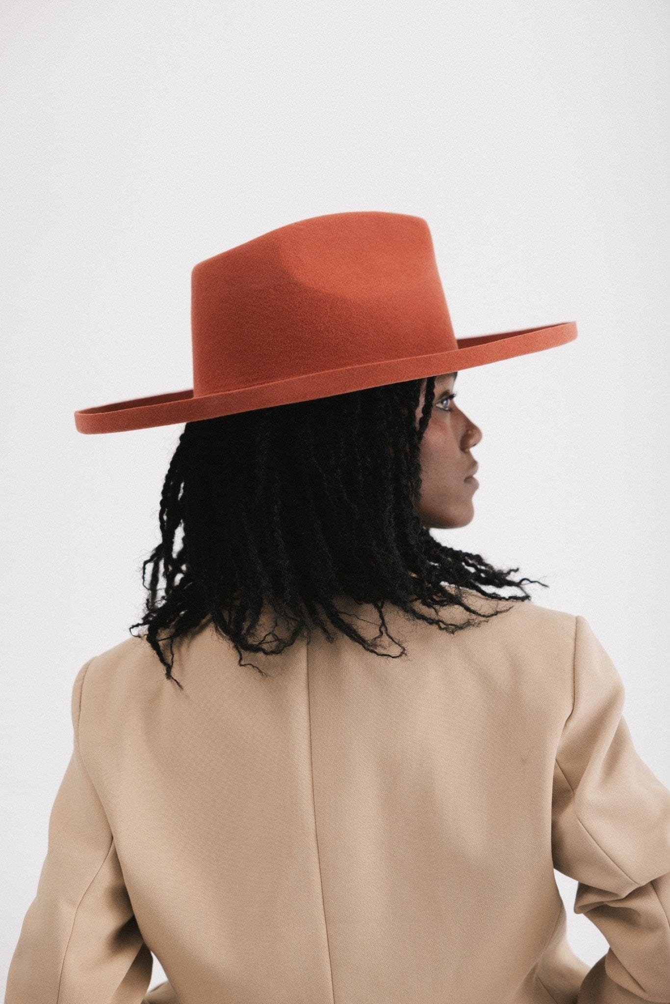 Woman wearing a clay colored pencil brim fedora hat with a wide brim and a light brown blazer, facing behind against a white background #color_clay