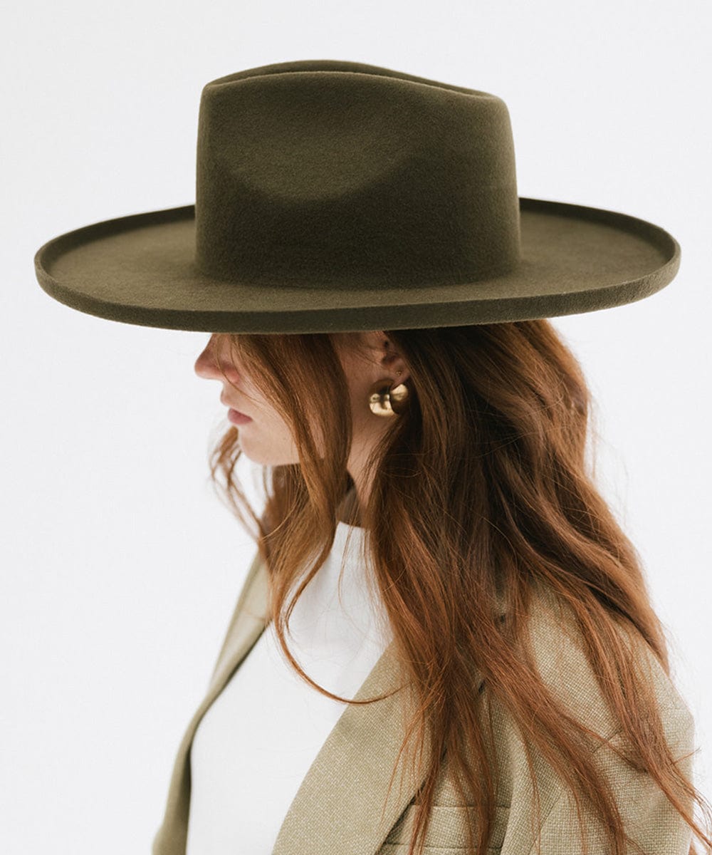 Woman wearing a moss colored pencil brim fedora hat with a wide brim and a white top with a beige blazer against a white background #color_limited edition moss