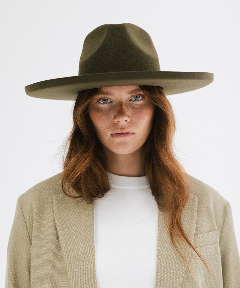 Woman wearing a moss colored pencil brim fedora hat with a wide brim and a white top with a beige blazer against a white background #color_limited edition moss