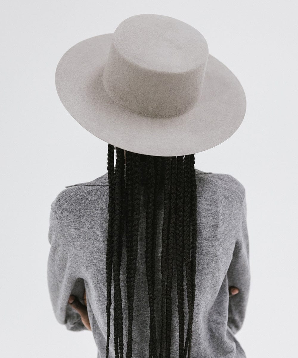 Woman with long hair wearing a light grey boater-style hat and a grey shirt, facing behind against a plain background #color_light grey