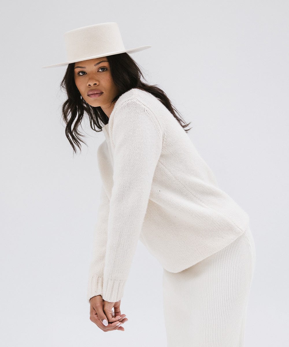 Woman wearing an off white boater-style hat, a white sweatshirt and white skirt, posing against a plain background #color_off white