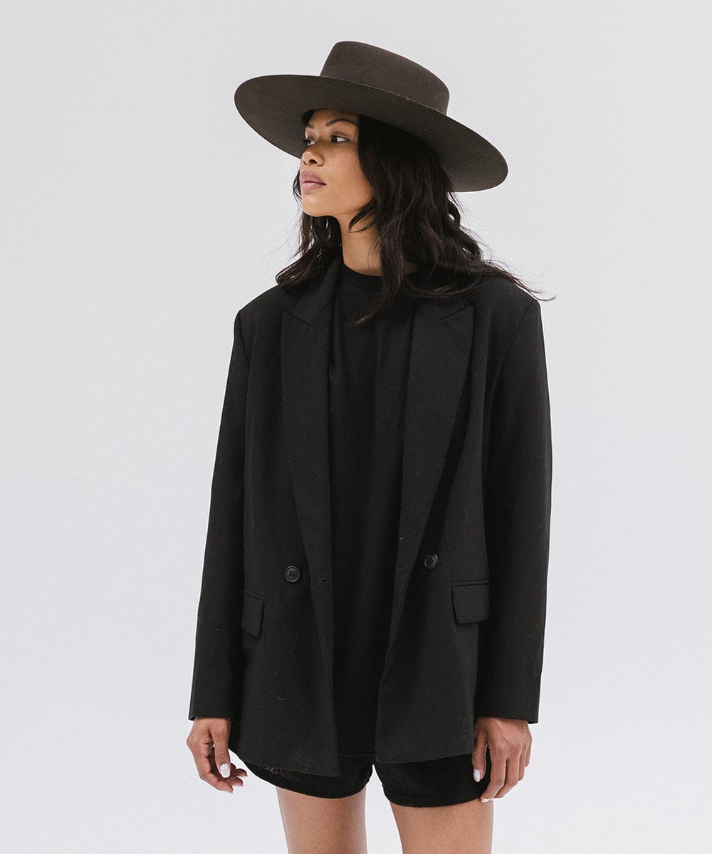 Woman wearing a dark brown wide brim felt hat with a triangle crown, a black t-shirt with a black blazer and black shorts, looking sideways against a white background #color_dark brown