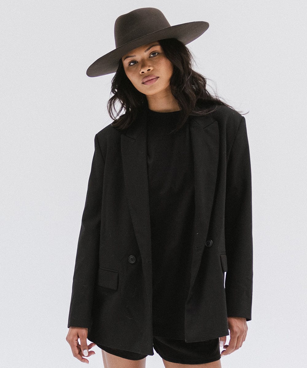 Woman wearing a dark brown wide brim felt hat with a triangle crown and a black t-shirt with a black blazer and black shorts against a white background #color_dark brown