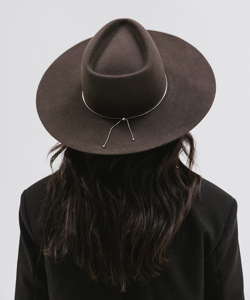 Woman wearing a dark brown wide brim felt hat with a triangle crown and a hat band, and a black blazer, facing behind against a white background #color_dark brown